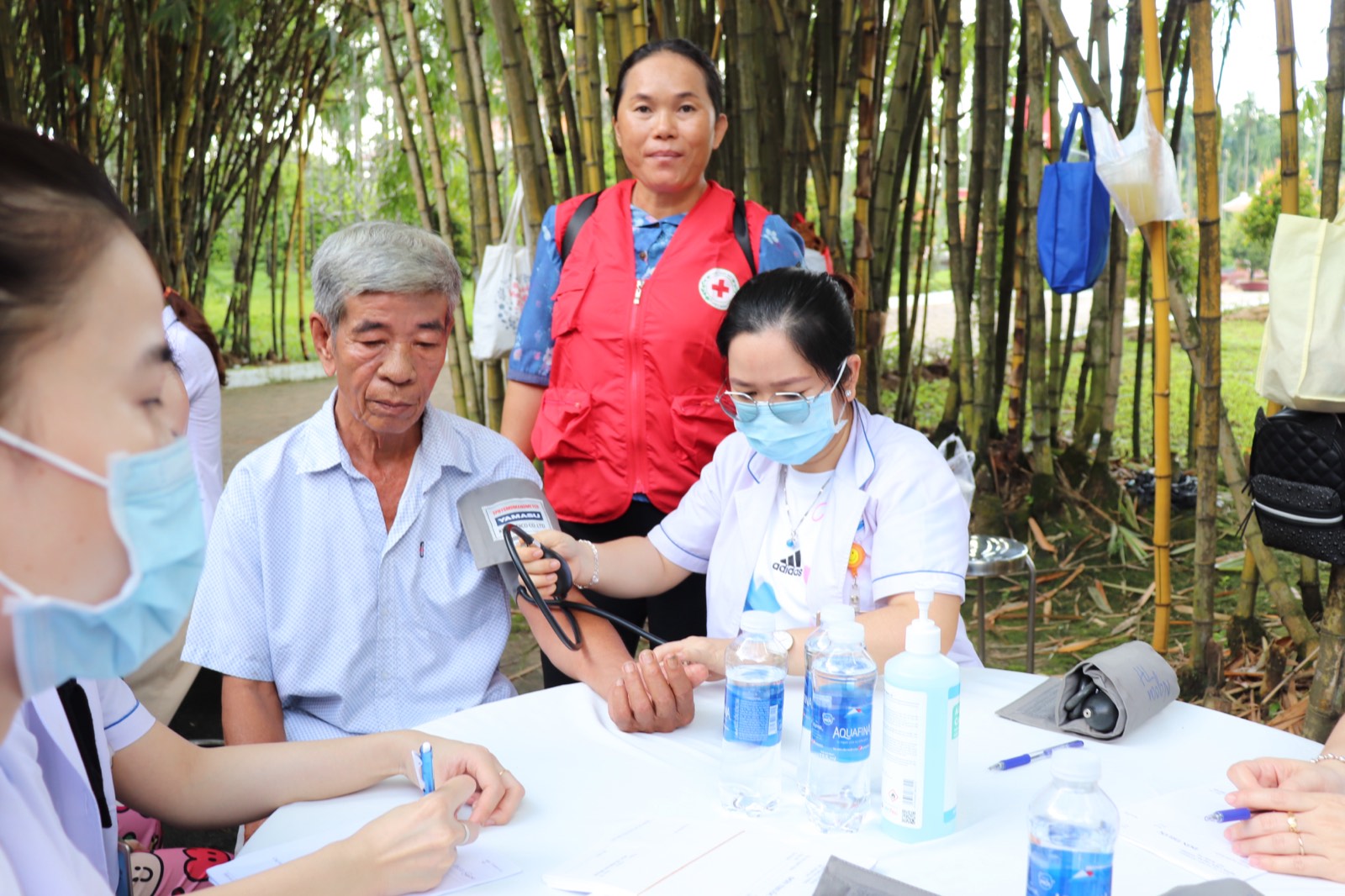 Điều dưỡng đo huyết áp cho một người cao tuổi tại bàn khám ngoài trời, tình nguyện viên Chữ thập đỏ đứng cạnh