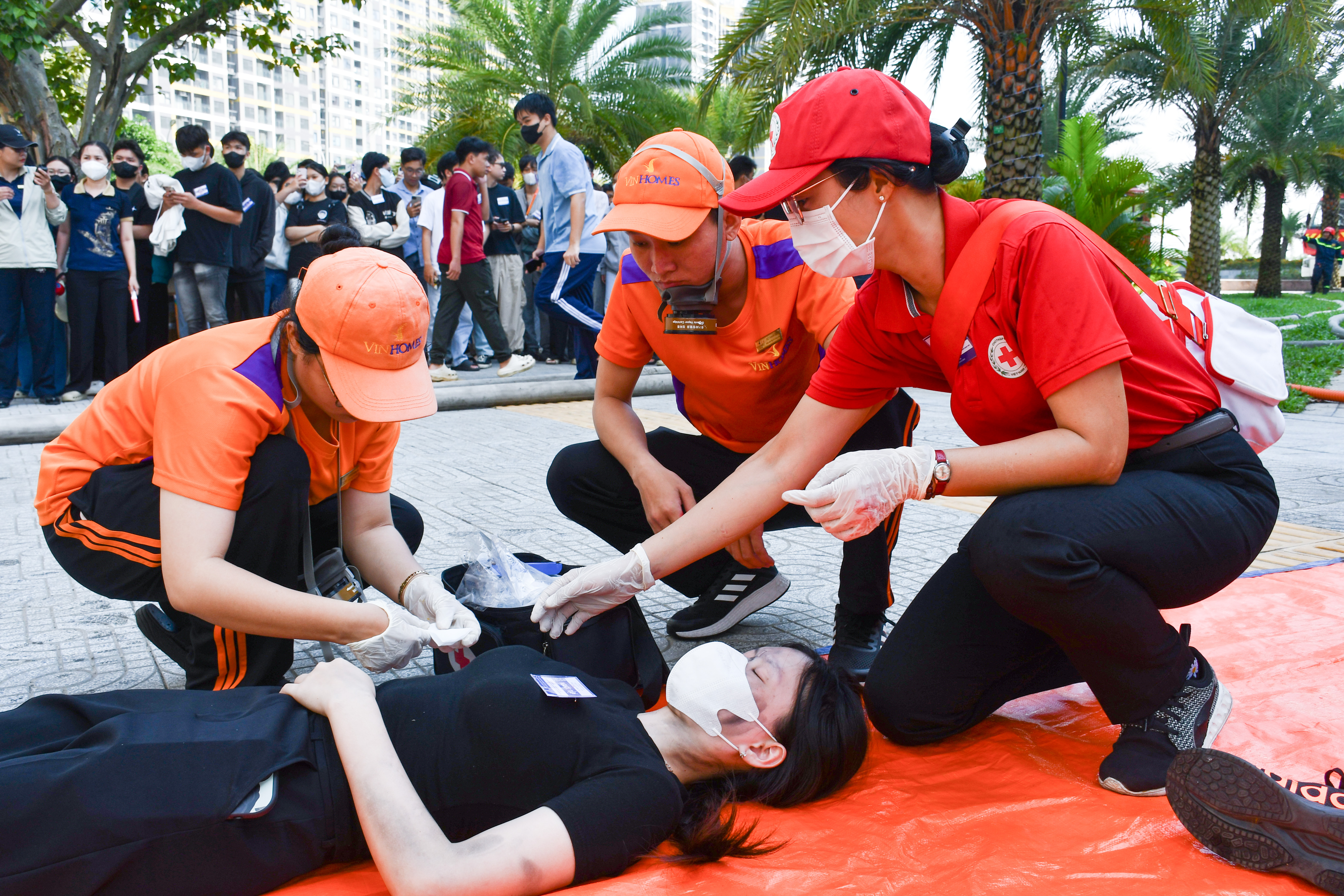 Cán bộ Hội Chữ thập đỏ hướng dẫn hai học viên thực hành sơ cấp cứu cho một người nằm trên tấm bạt trong buổi diễn tập ngoài trời